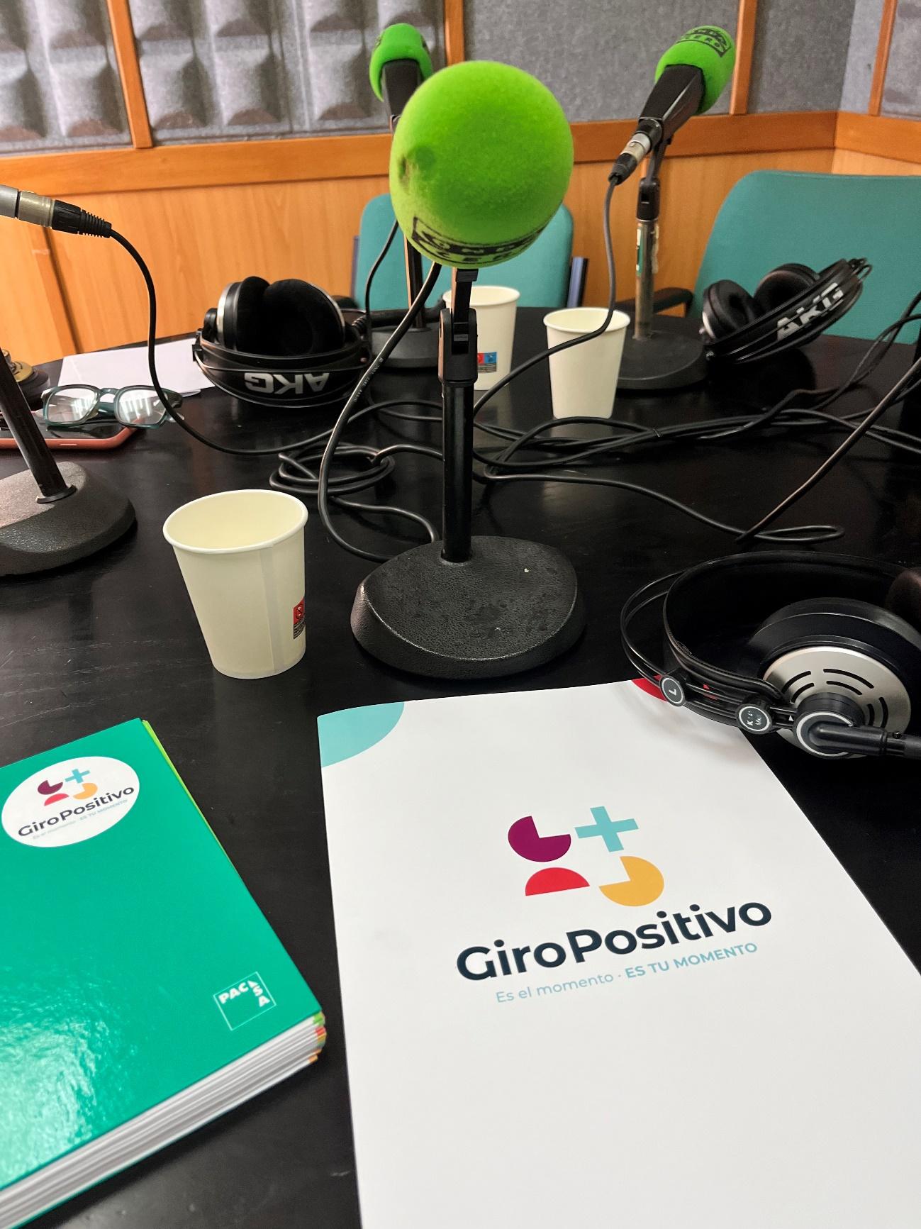 mesa de estudio de radio con micrófonos, libretas y voli, cascos y un vaso de agua.